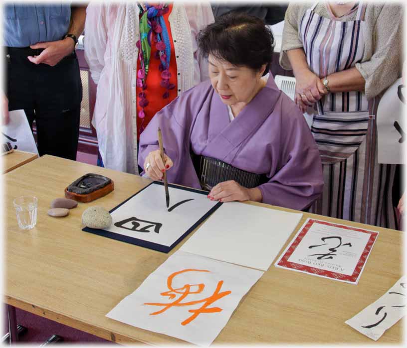 Six calligraphy class members standing around seated woman writing with brush.