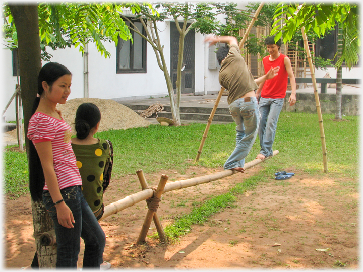 Two students trying to walk on a pole held at one end by a rope, two watchers.