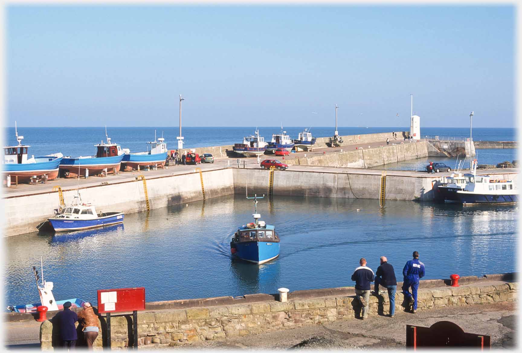 Two groups of men standing by harbour wall watching boat turning.