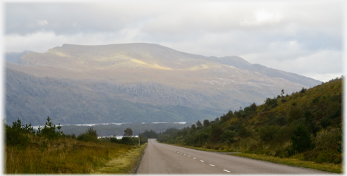 Road running down towards loch with wide flat hill beyond.