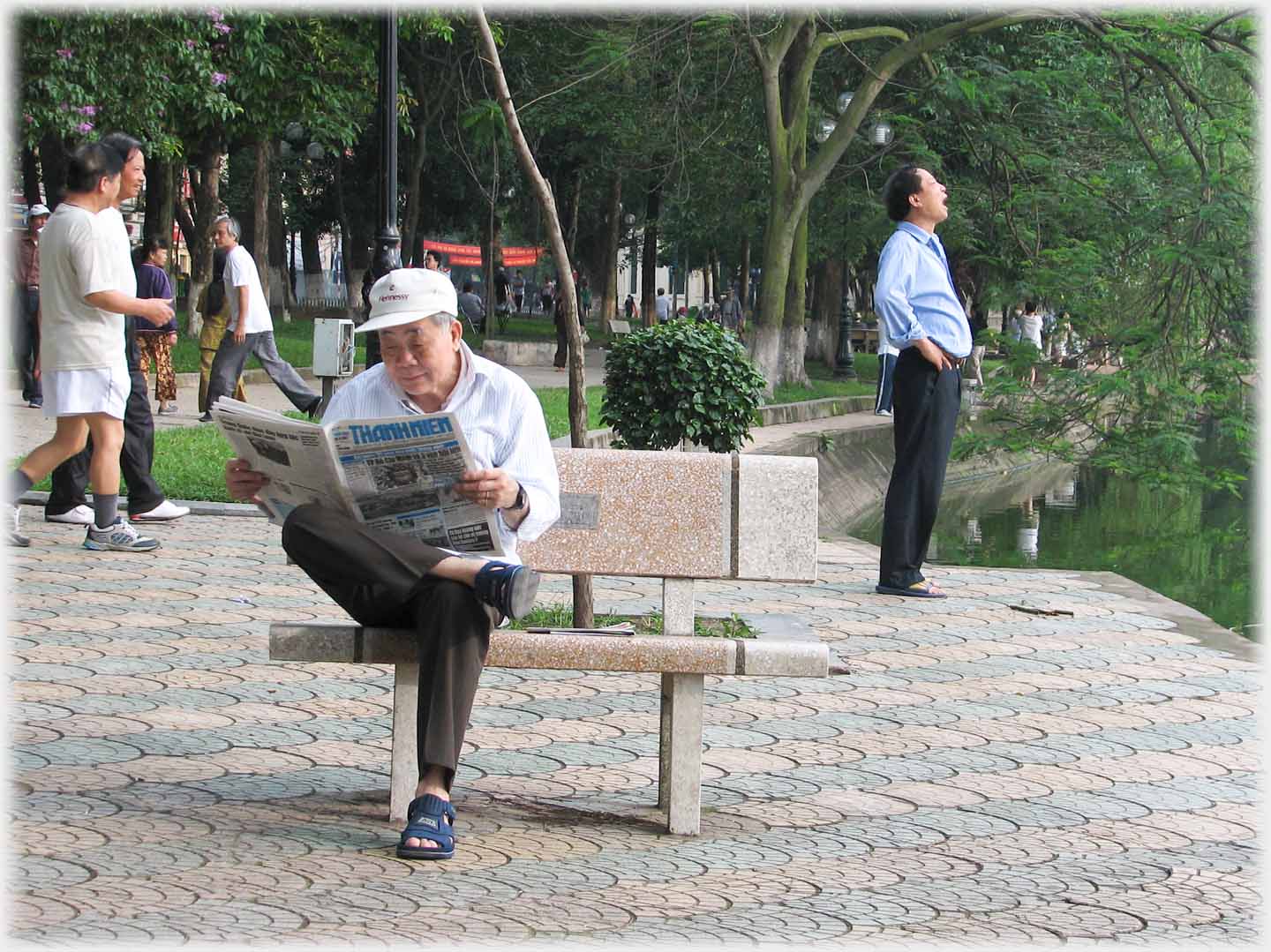 Man sitting on park bench reading newspaper, others behind him.