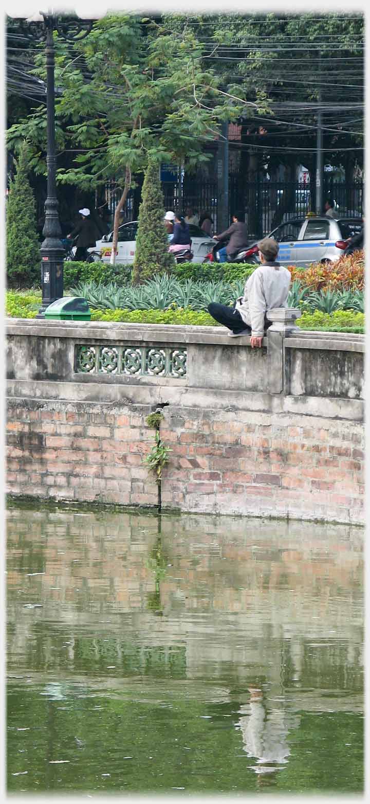 Man squatting on a parapit leaning against a small pillar, some feet above water.