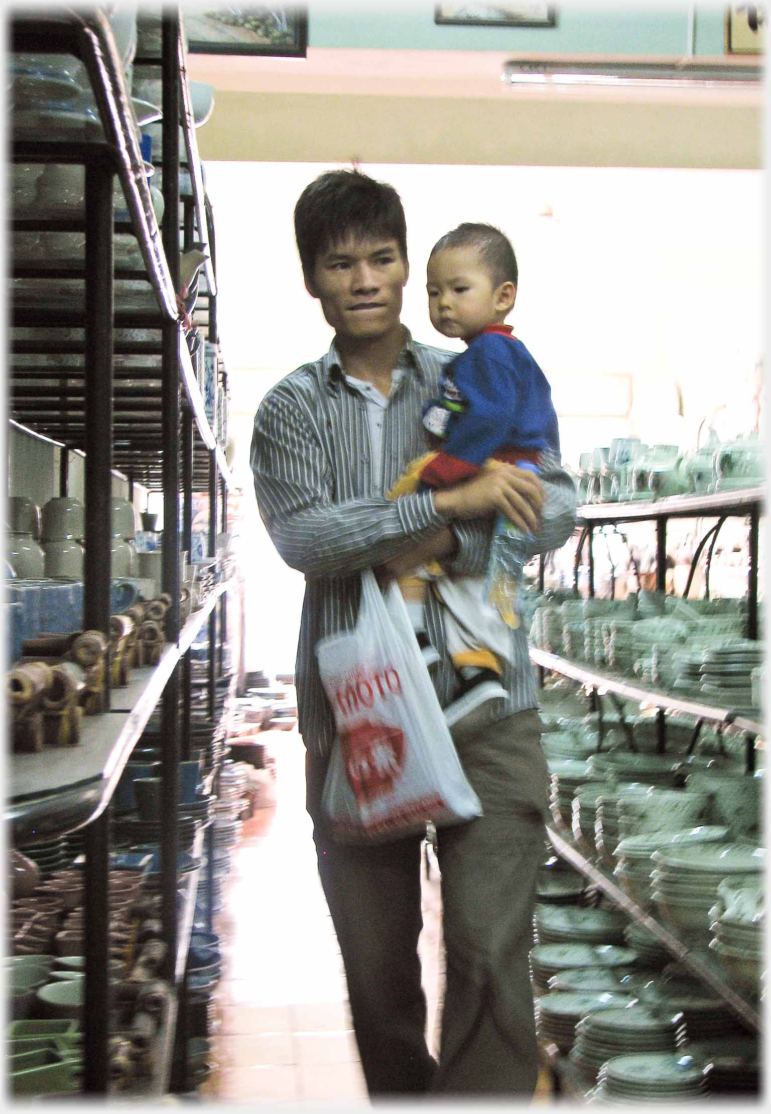 Man holding infant walking between shelves of ceramics.