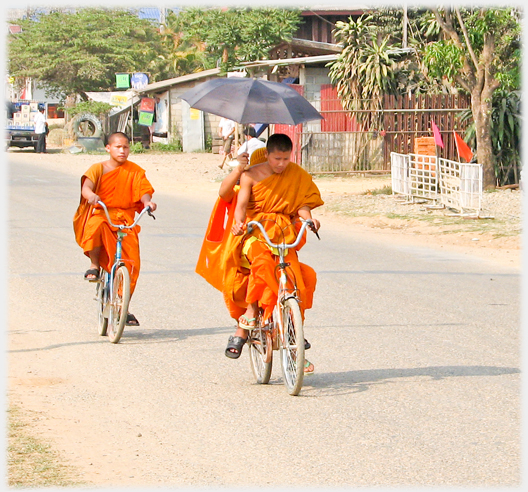 Two monks on a bicycle the passenger holding an umbrella, another monk close behind.