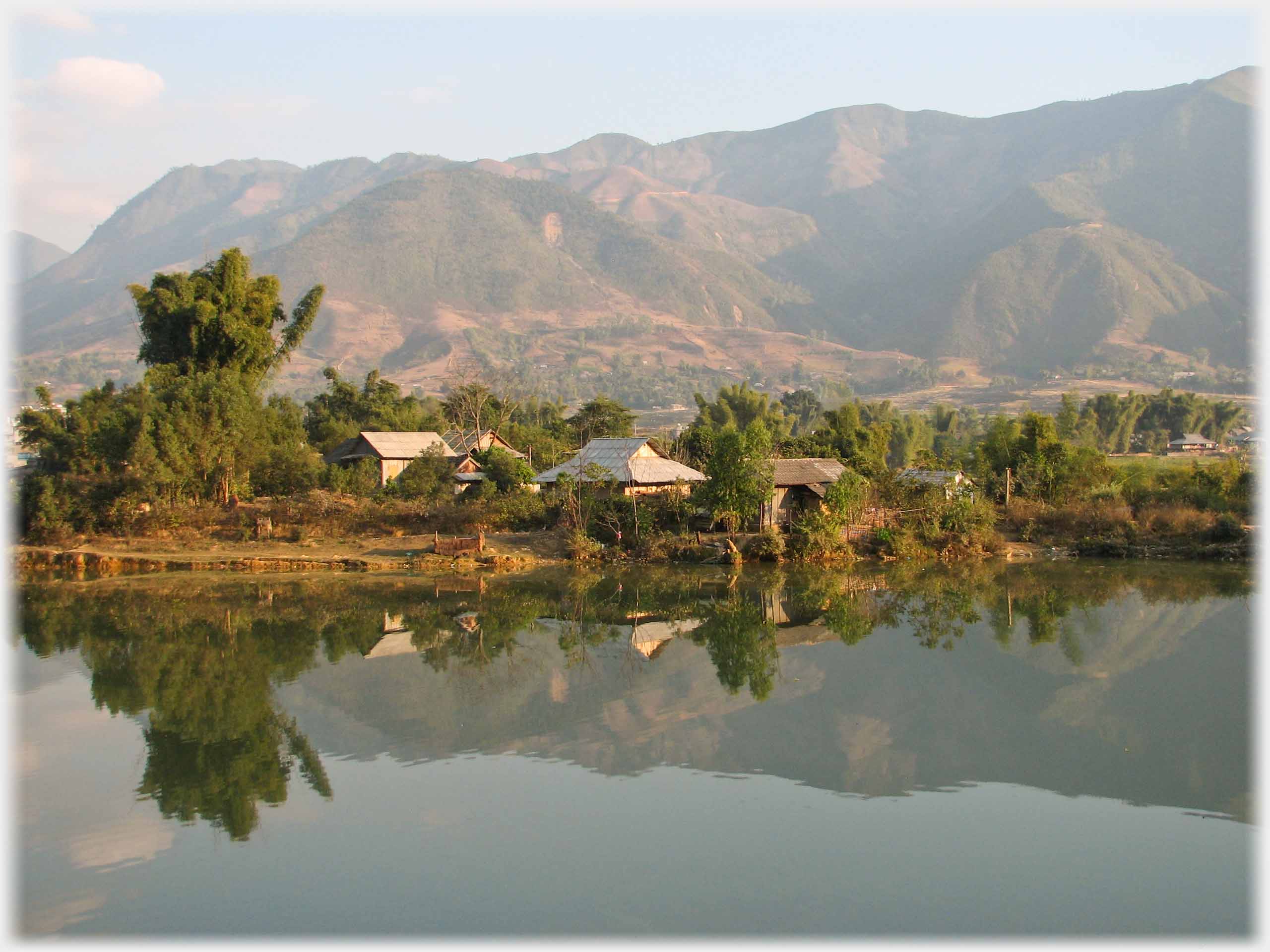 Houses clustered by river with bamboos and mountains beyond.