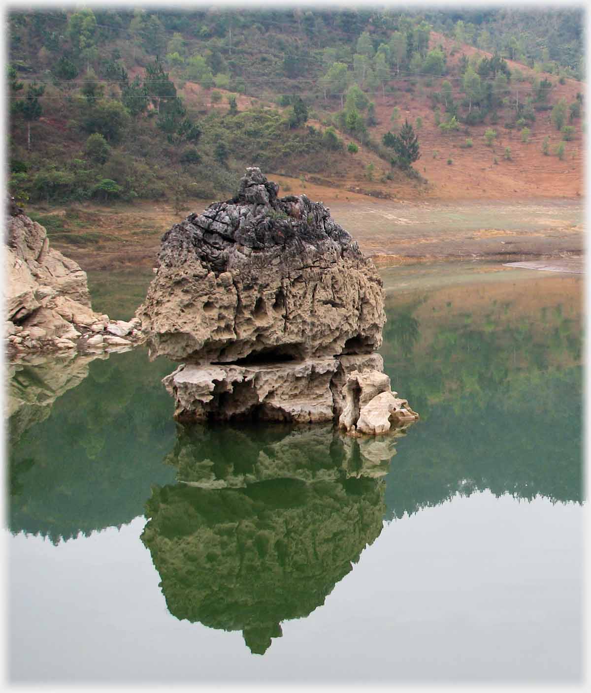 Much pitted rock standing in river with reflection.