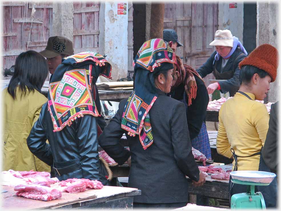 Two women with backs to camera at meat stall, both in black with elaborate coloured headdresses.