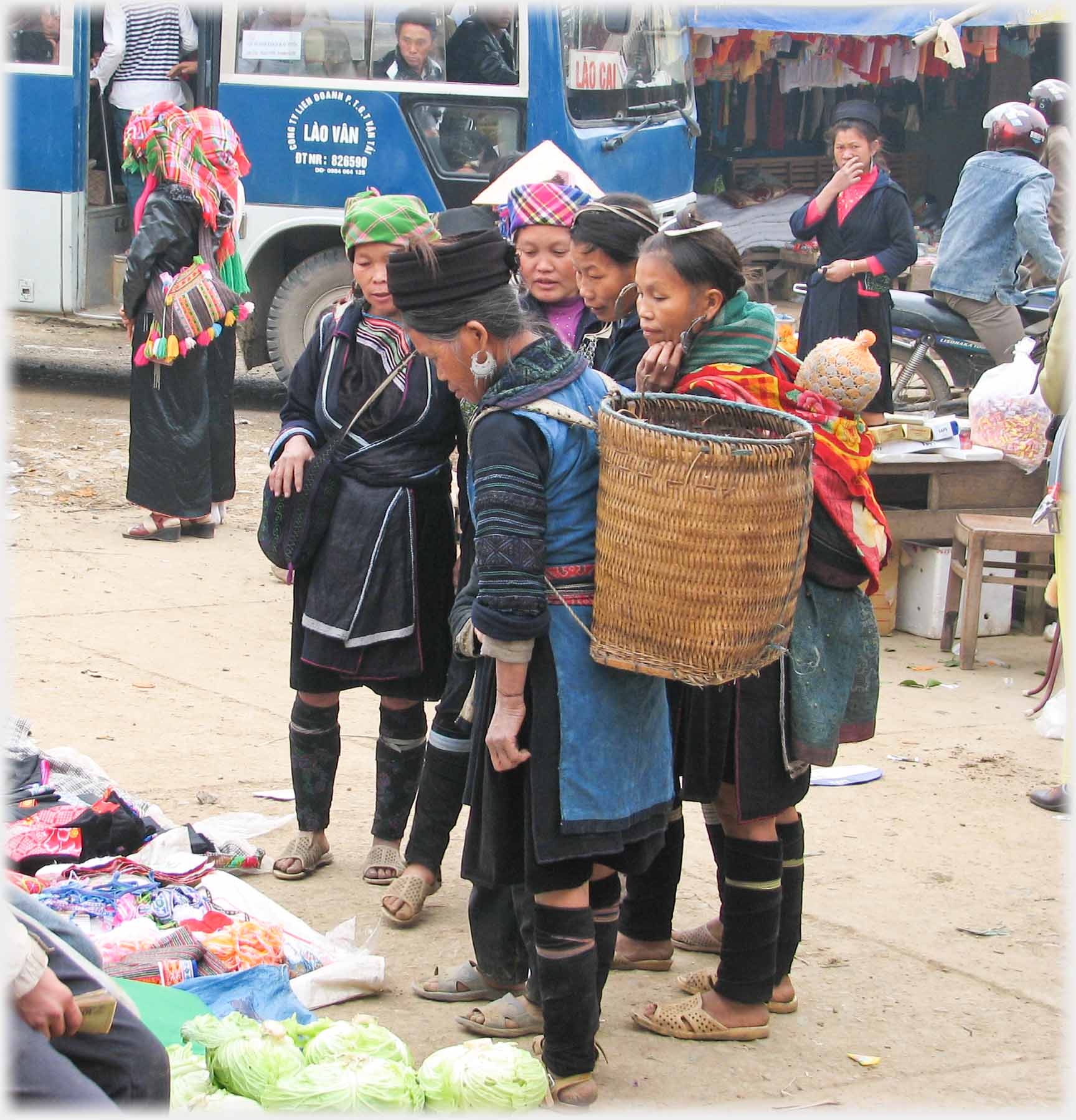 Group of women in black outfits including socks, knees bare, looking at produce, nearest one with large cylindrical woven basket on her back.