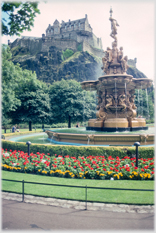 Large ornate fountain with flower beds and castle on rock behind.
