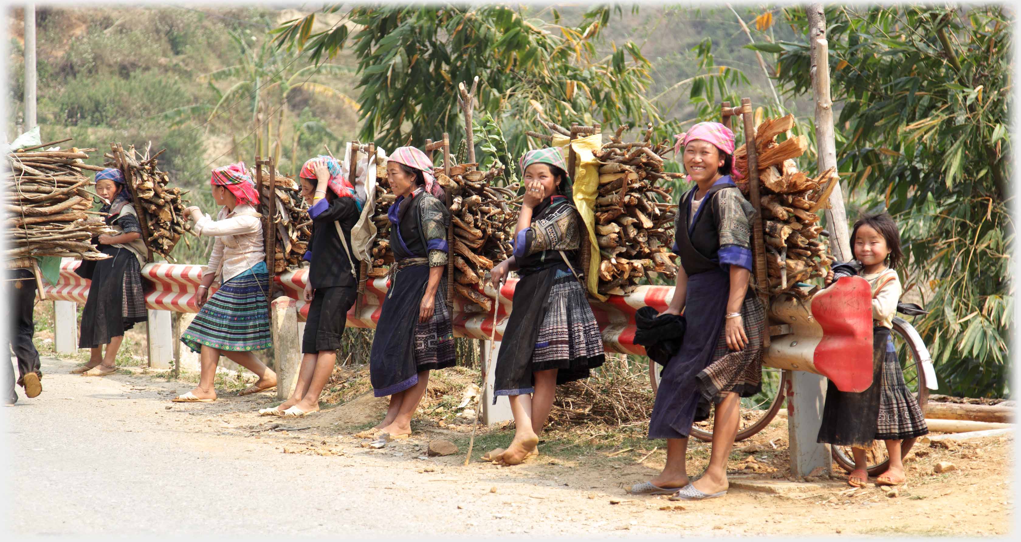 Row of six women with large packs of firewood on their backs, resting them on a crash barrier.