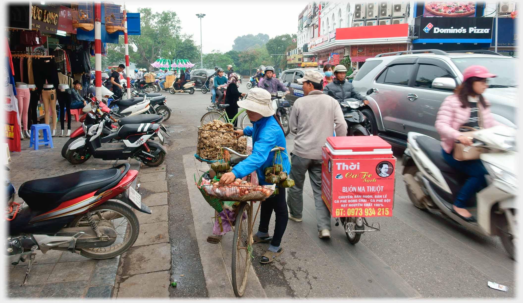 Woman with baskets of ginger and onions on a bicycle on a busy street.