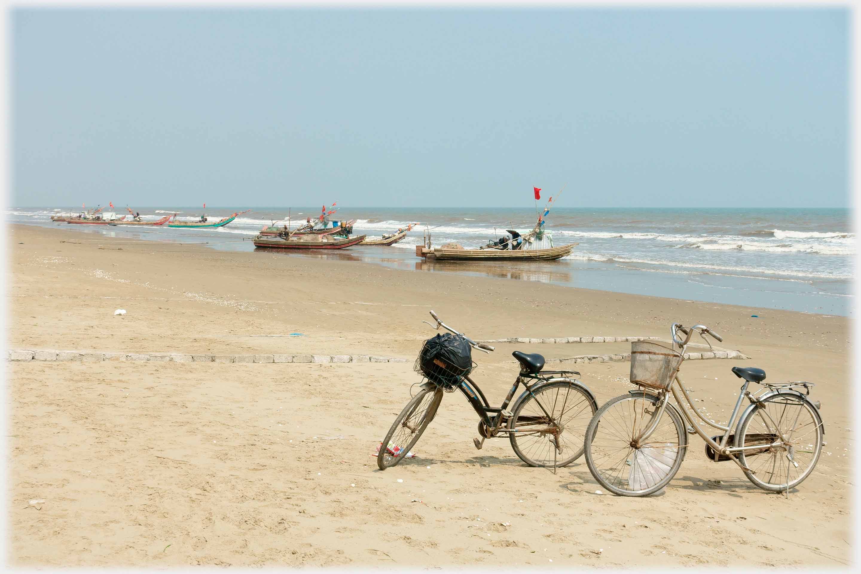 Two cycles parked on the beach, beached boats in the background.