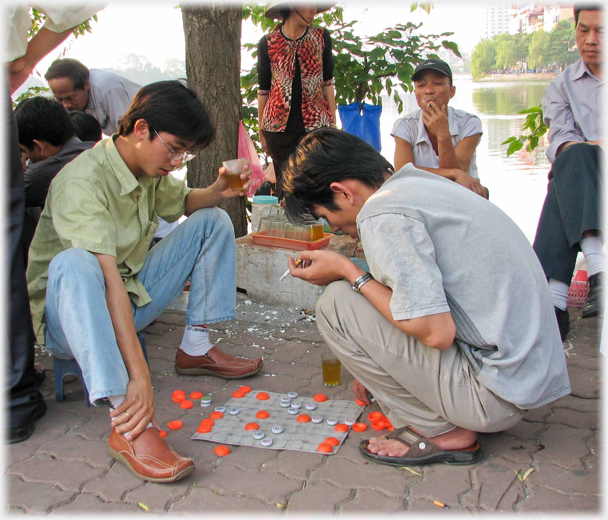 Two men, one with tea and one with cigarette are playing, watched from further off by two men, woman selling tea looking away.