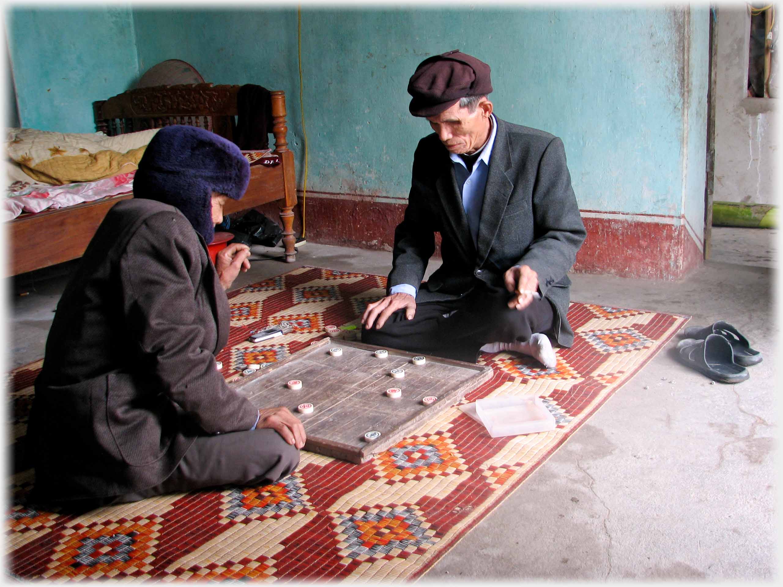 Two men cross-legged on floor, a board with pieces between them.