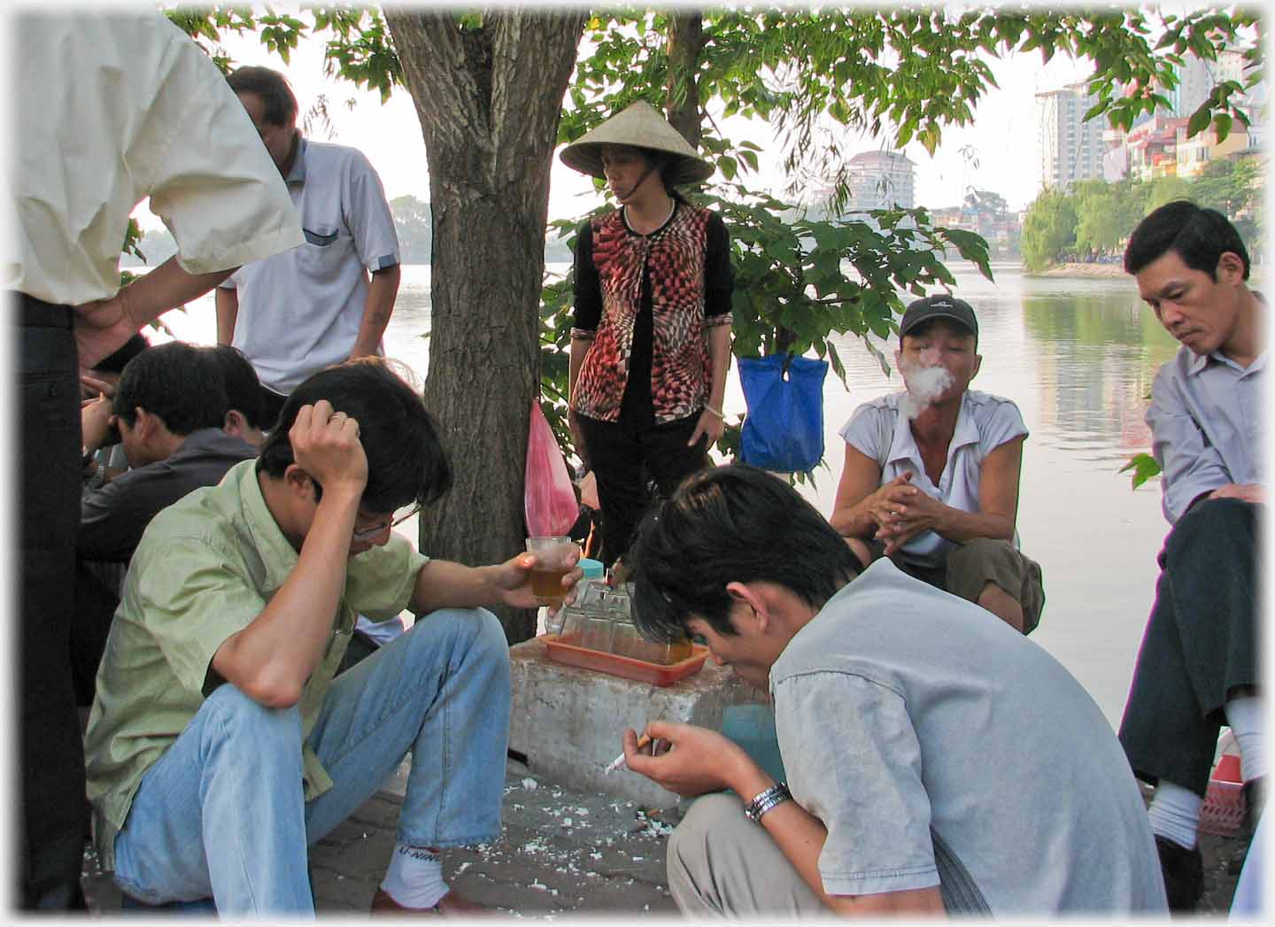 Chess players looking down, holding tea and hoolding cigarette. Tea seller behind. Man blowing smoke.