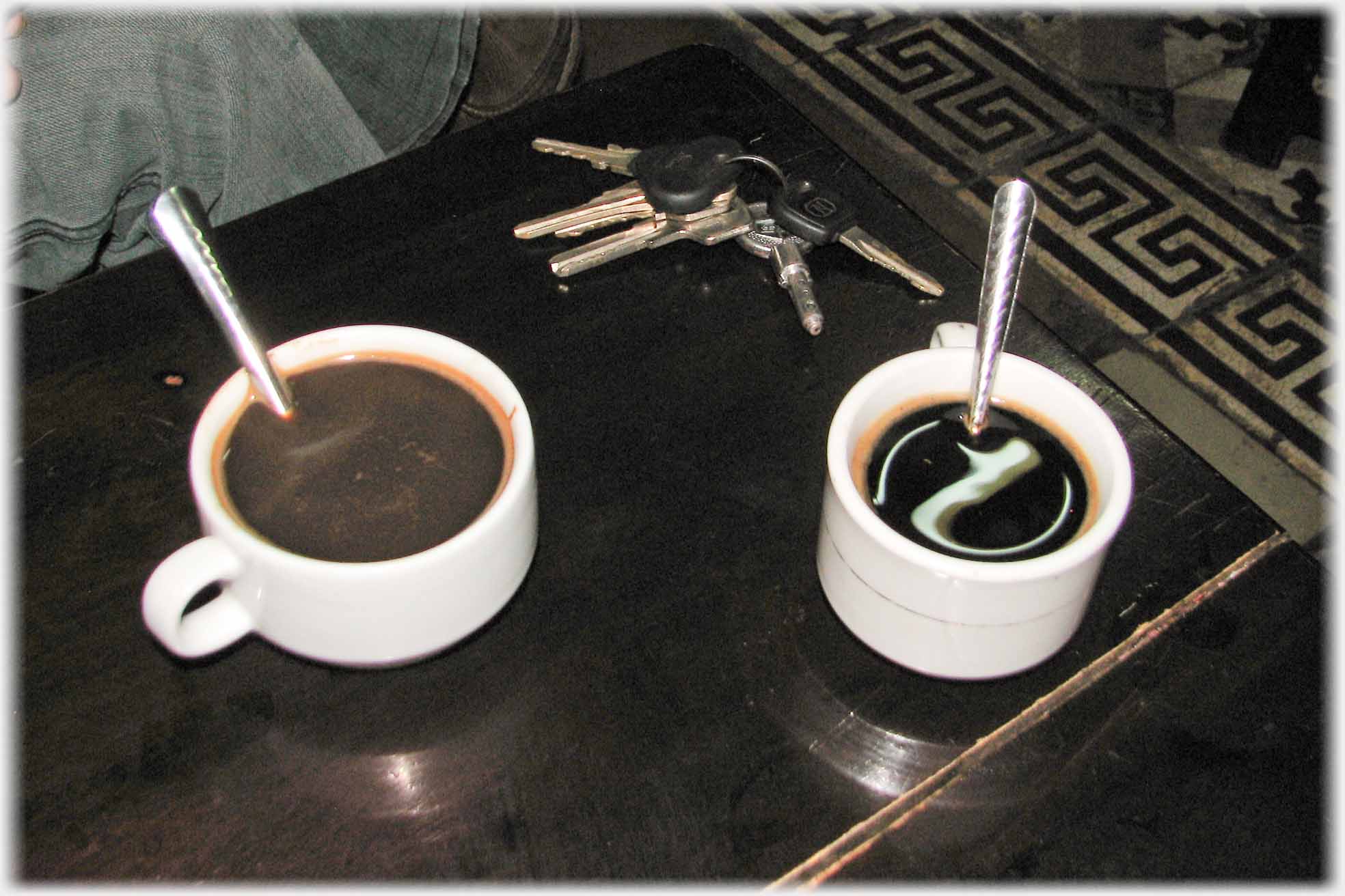 Two small coffee cups with spoons in them, and keys on table.
