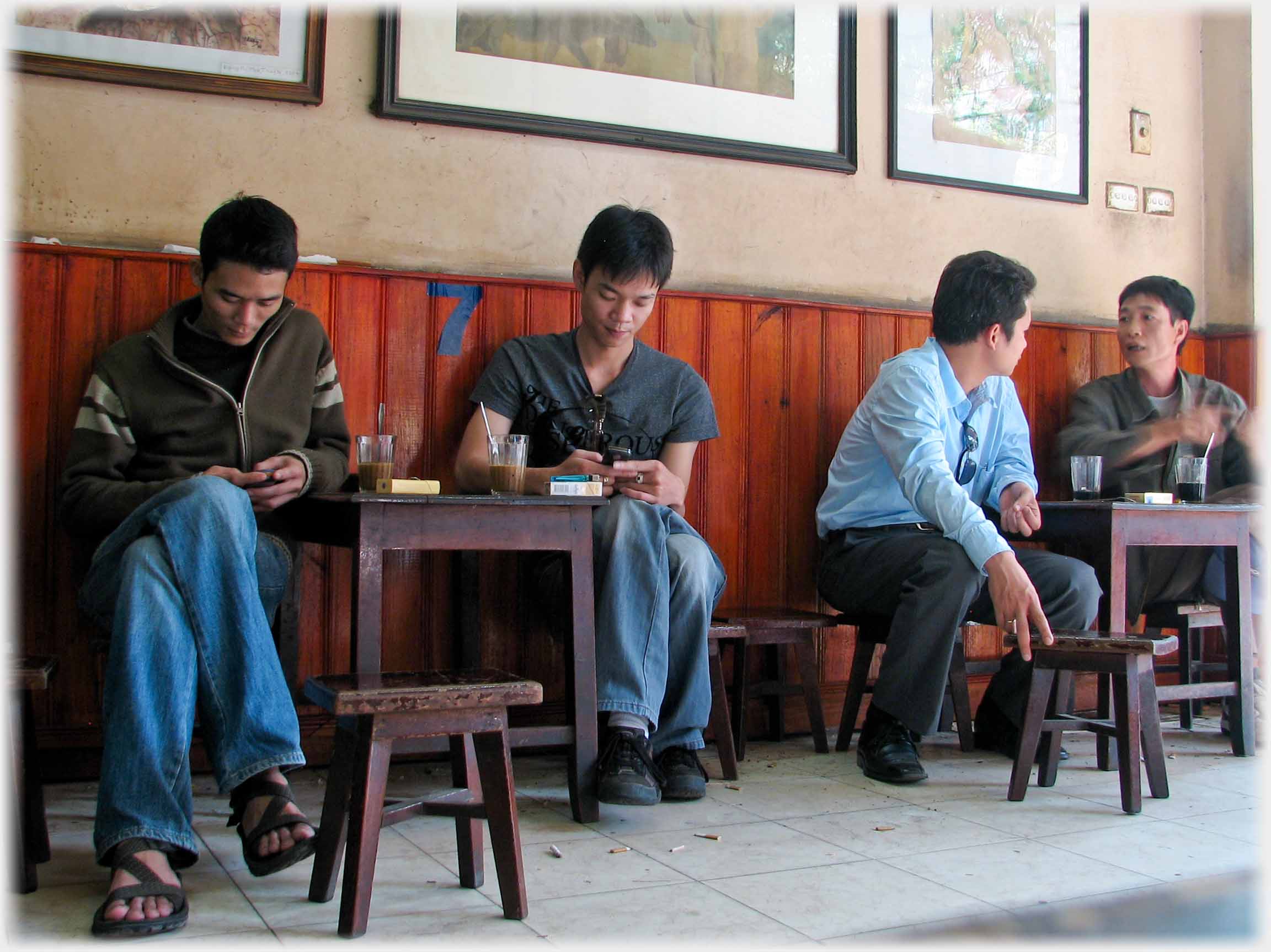 Two men together at a table separately texting, two further men at another table conversing.