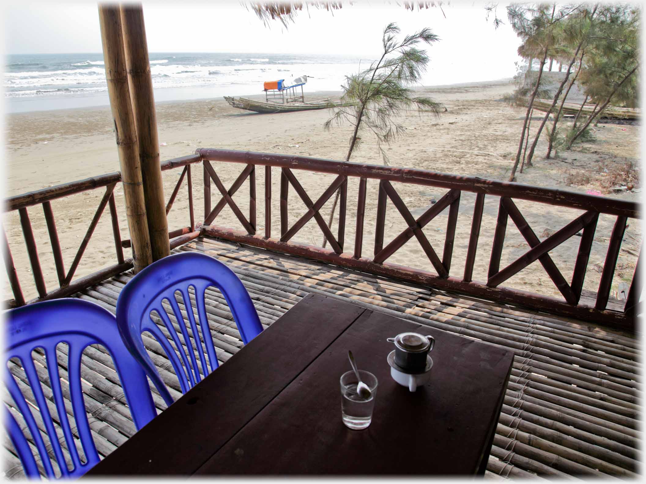Coffee filtering into a cup with glass of water on table, on veranda, next to beach, boat and sea.