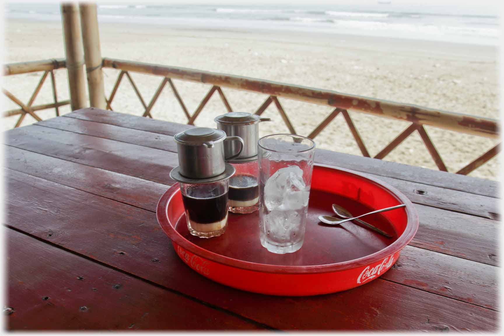 Two coffee glasses with filters sitting on them beside a glass of ice on table on veranda by beach.