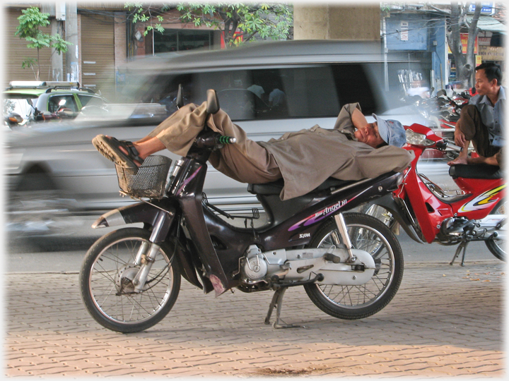Man sleeping on motorbike, another man sitting on his, and traffic passing close by.