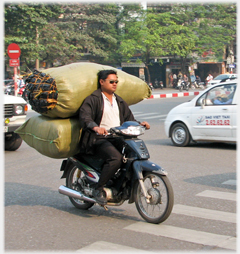 Man wearing sunglasses, no helmet or mask, with two large sacks as pillion.