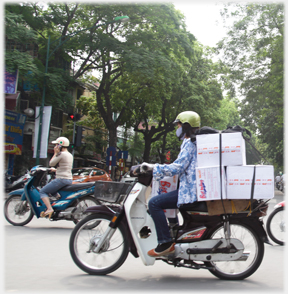 Two helmeted riders, woman with boxes, man phoning.