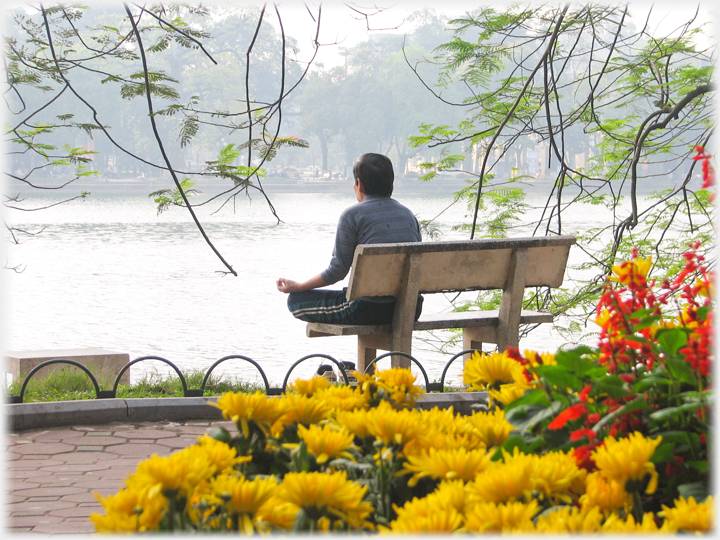 Man on seat by lake meditating, flowers in foreground.