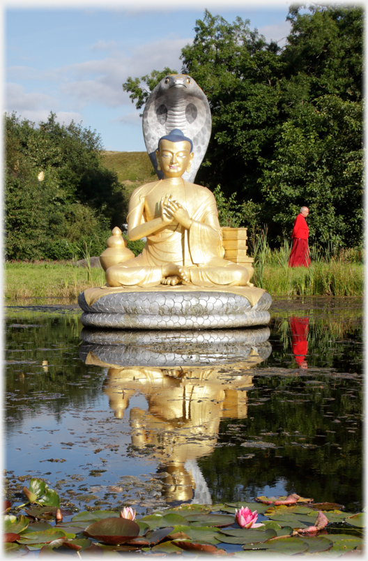 Golden sitting statue in pool, with monk walking past and waterlily in foreground.