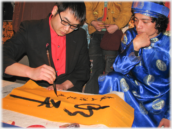 Man in blue robes watching male calligrapher.