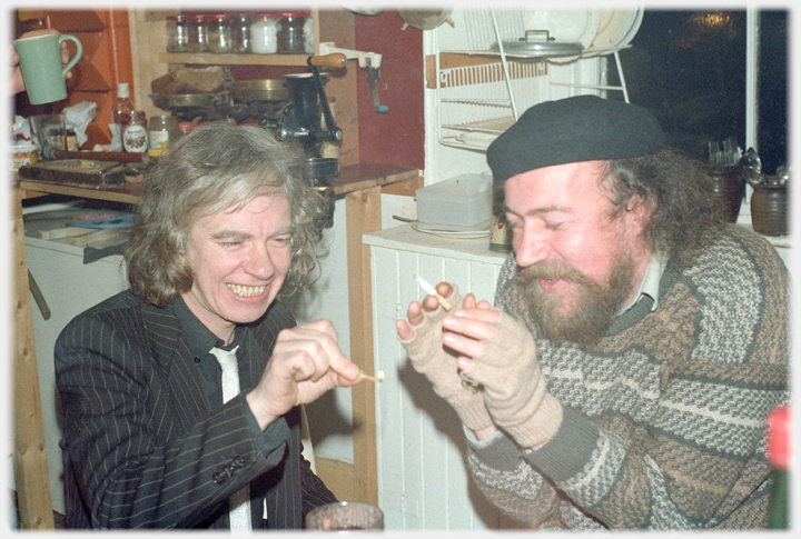 Two men, one holding a lit match the other warming his hands by it.