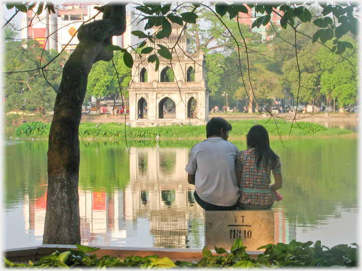 Couple sitting by lake with monument on island.