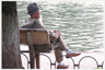 Man with beard sitting on bench, knees clasped, by water.