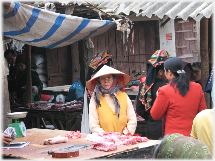 Woman in hat with large glasses by meat stall.