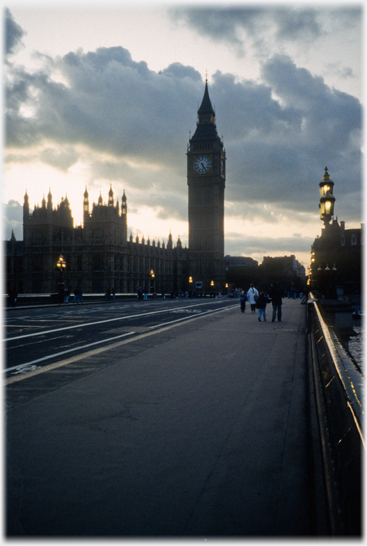 Big Ben clock tower viewed across a road, dark clouds and lamps.