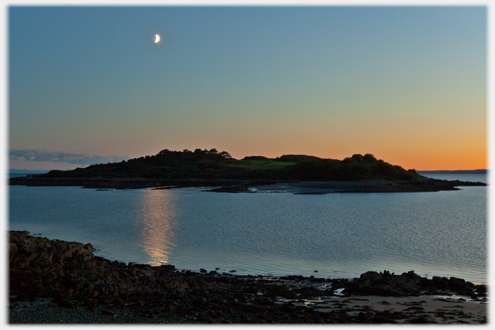 Moon in sky over sunset behind island.