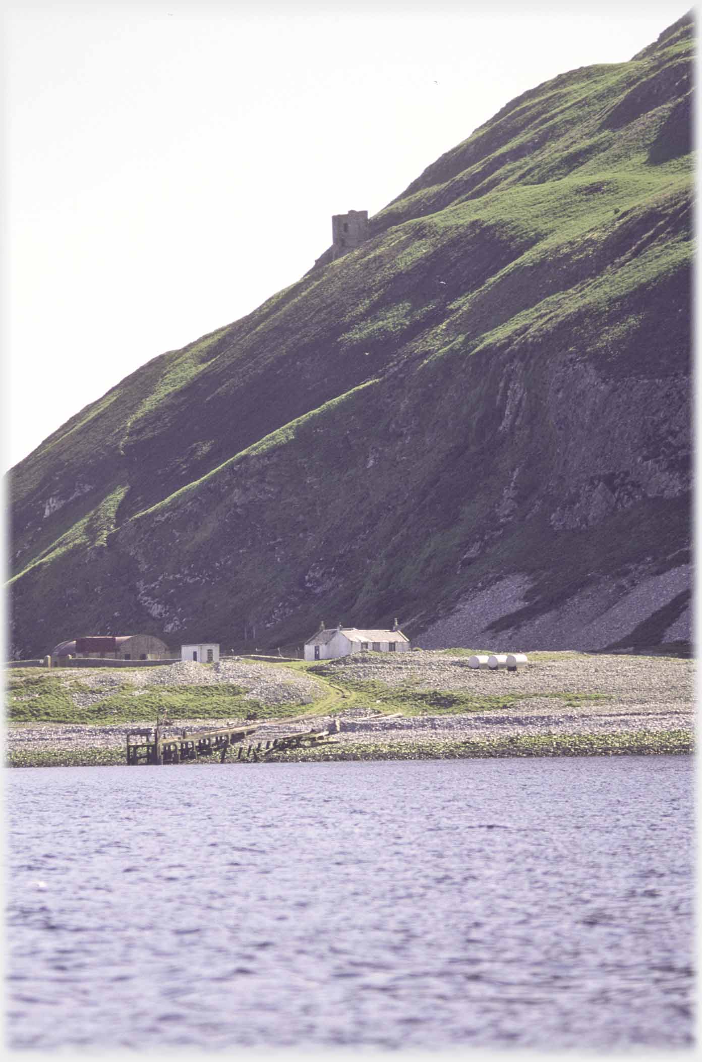 Steep hillside above houses by shore.