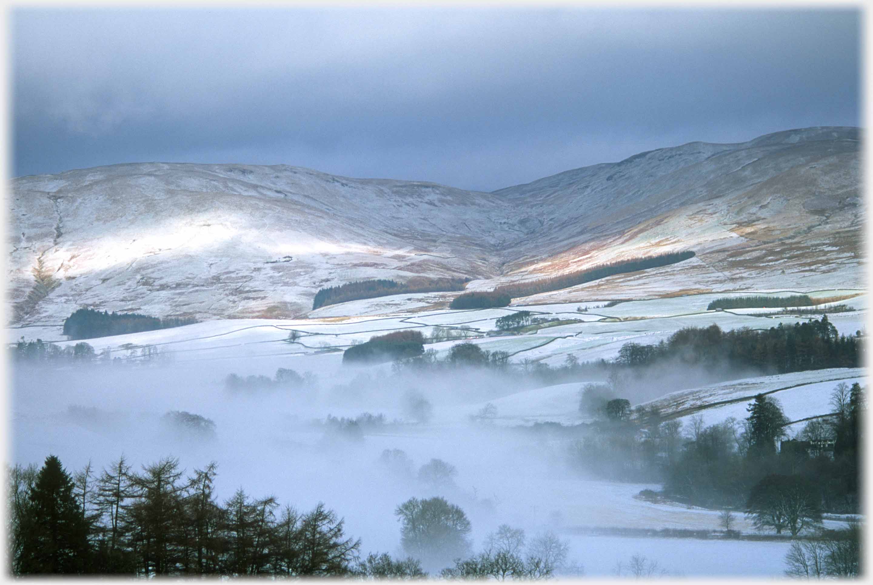 Fields and Treed valley with mist hanging and brackened hills beyond in light snow.