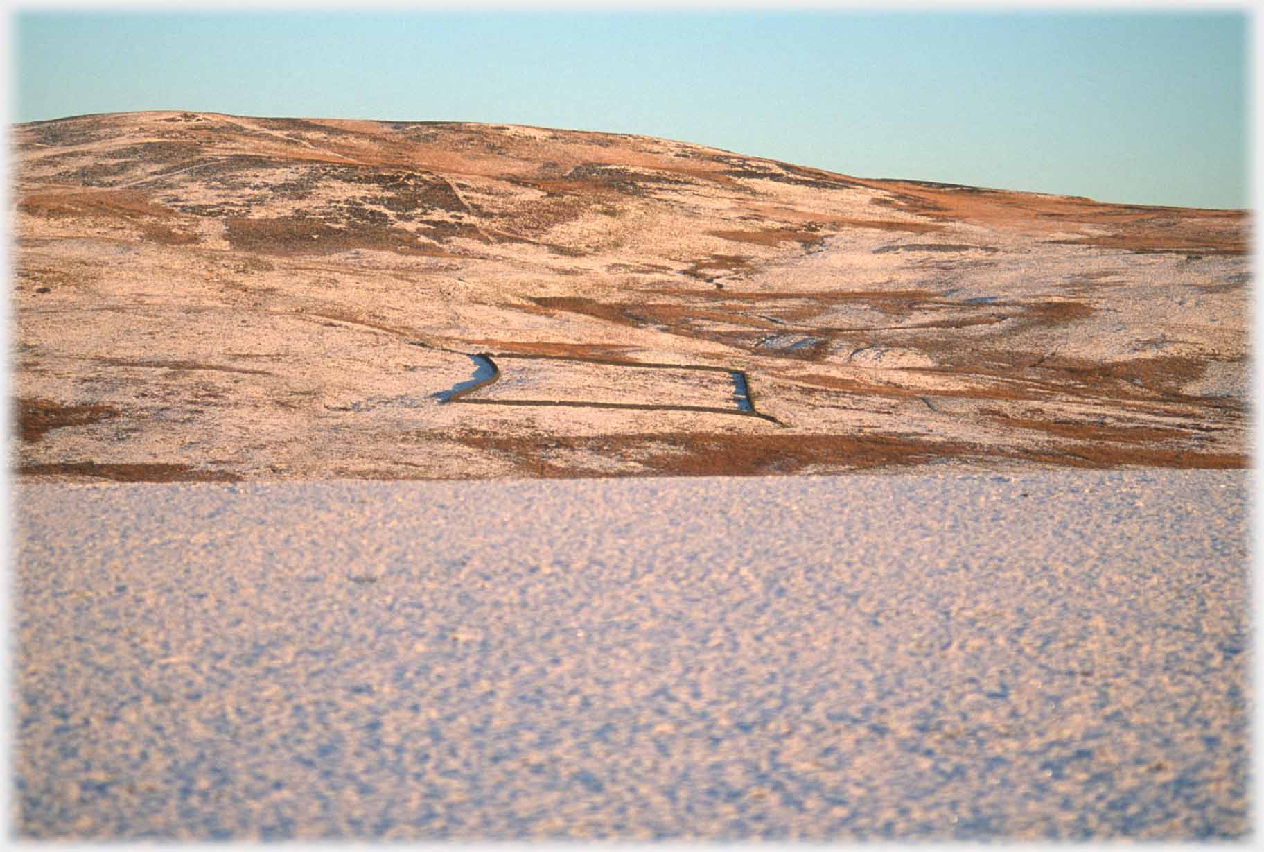 Large square dyke on snow sprinkled hillside.