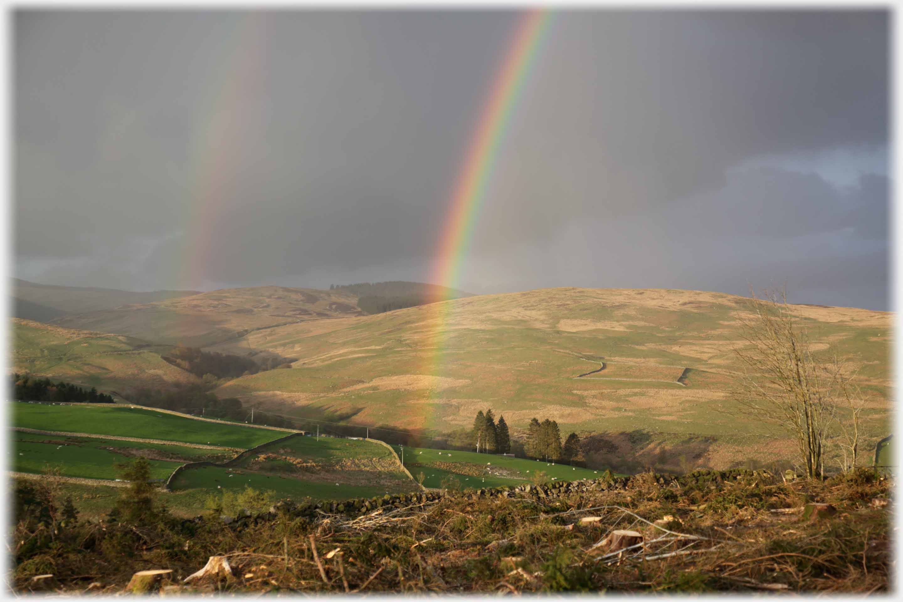 Black cloouds over hillsides with stong rainbow and its secondary just showing.