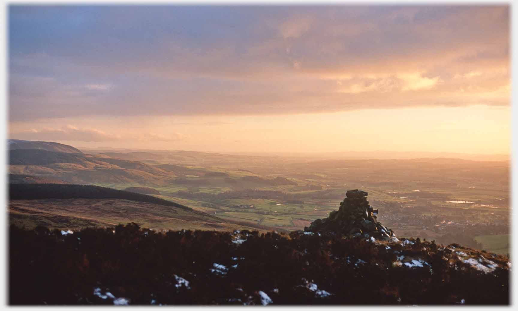 Cairn on hilltop with valley in sunset beyond.