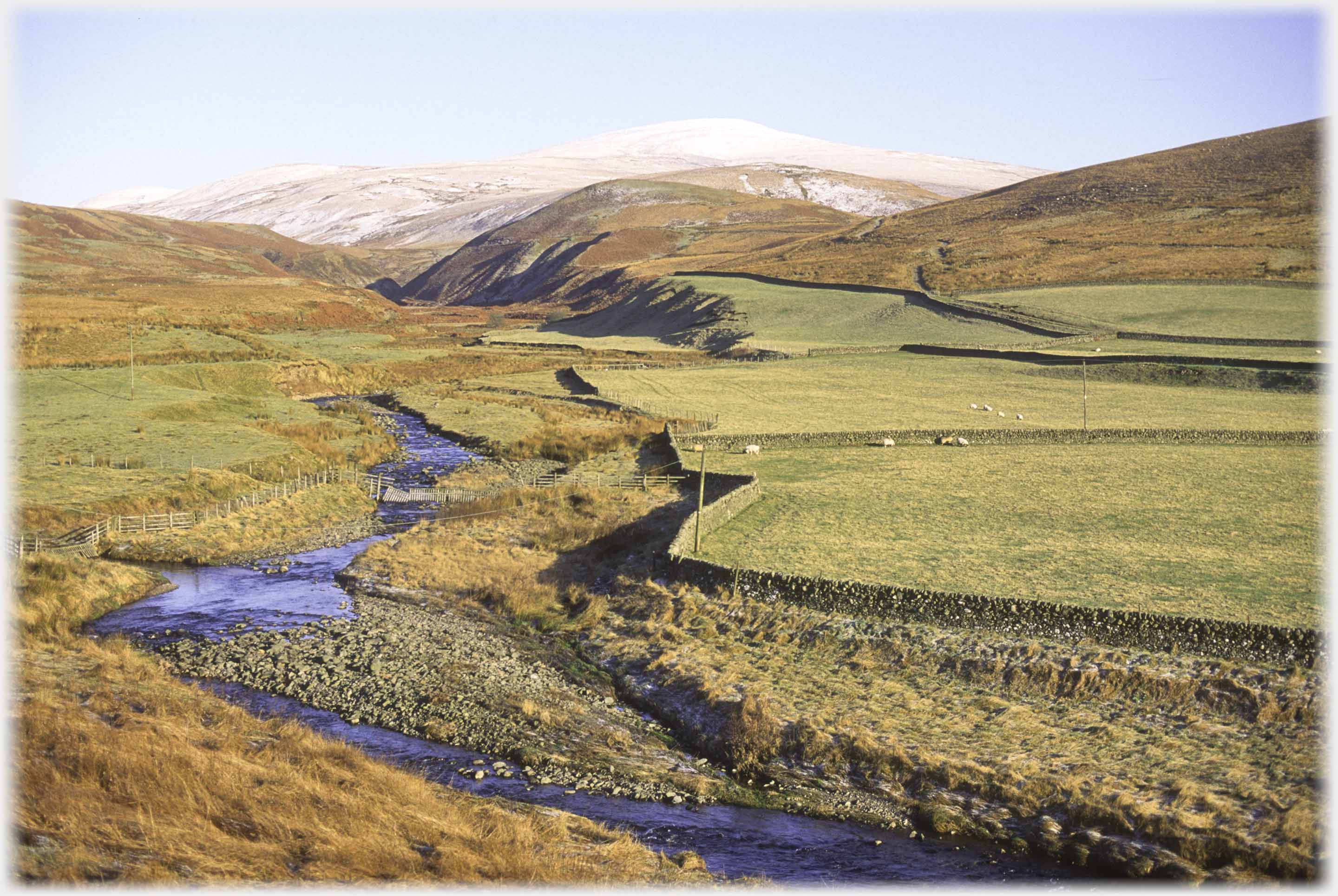 Fields and dykes by stream with snow covered hill in background.