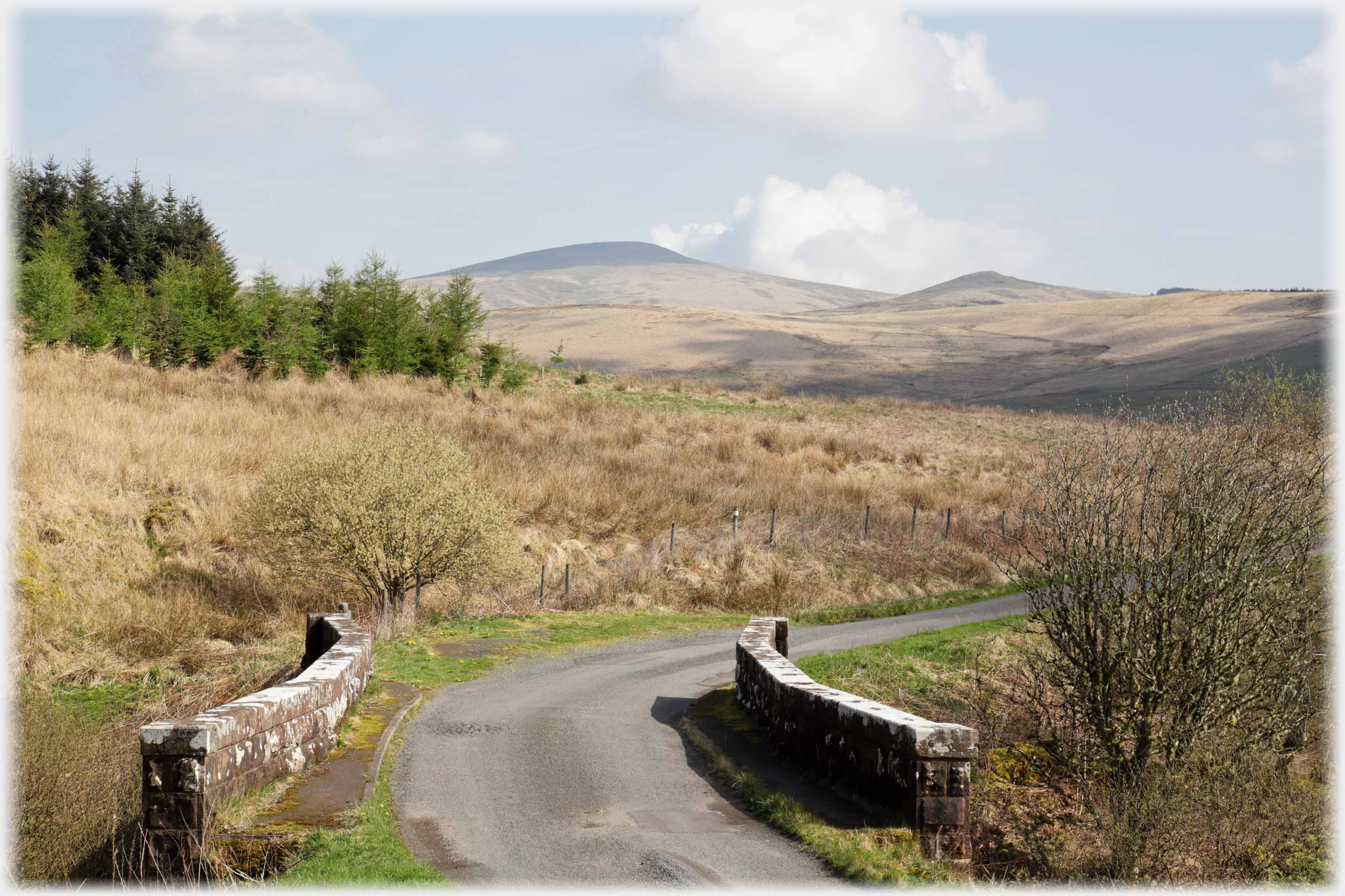 Road running over parapetted bridge pointing at hill.