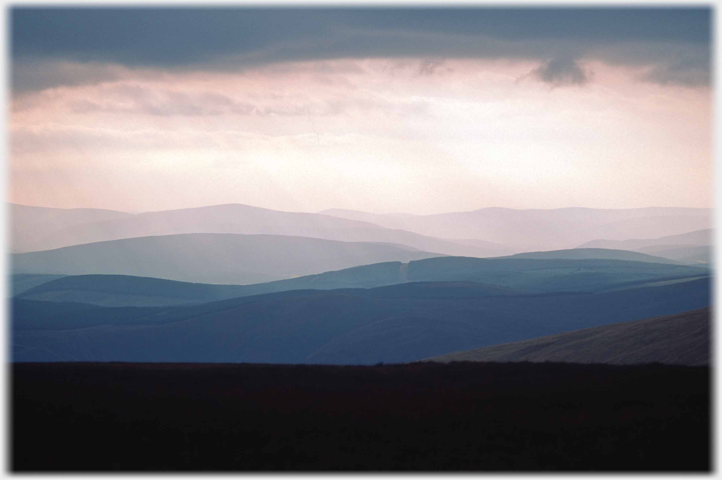 Looking out across range on range of hills in pink light and heavy cloud.