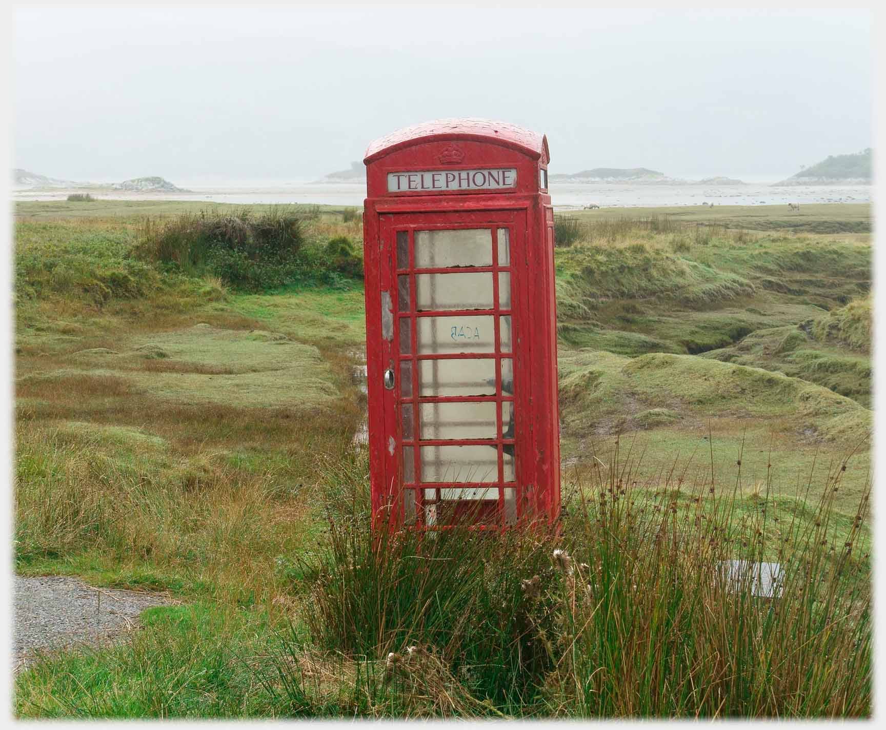 Tilting phone box with unkempt land and shore around.