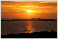 Deep orange sky with sinking sun reflected across water through island.