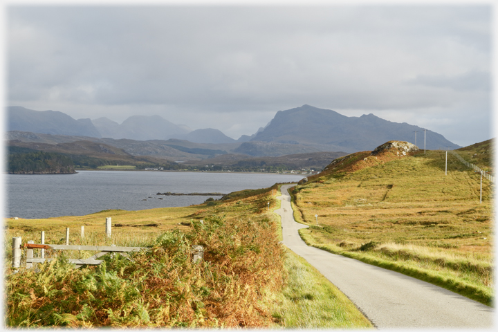 Single track road pointing at hills beyond houses on shore of loch.