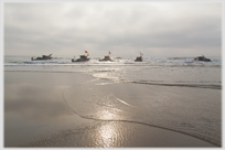 Five fishing boats in line at sea with shallow water waves in foreground.