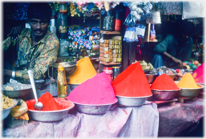 Steeply piled bowls of dyes along stall edge with man behind.