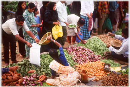 Vegetable sellers on ground piles of produce in front, woman filling a man's bag.