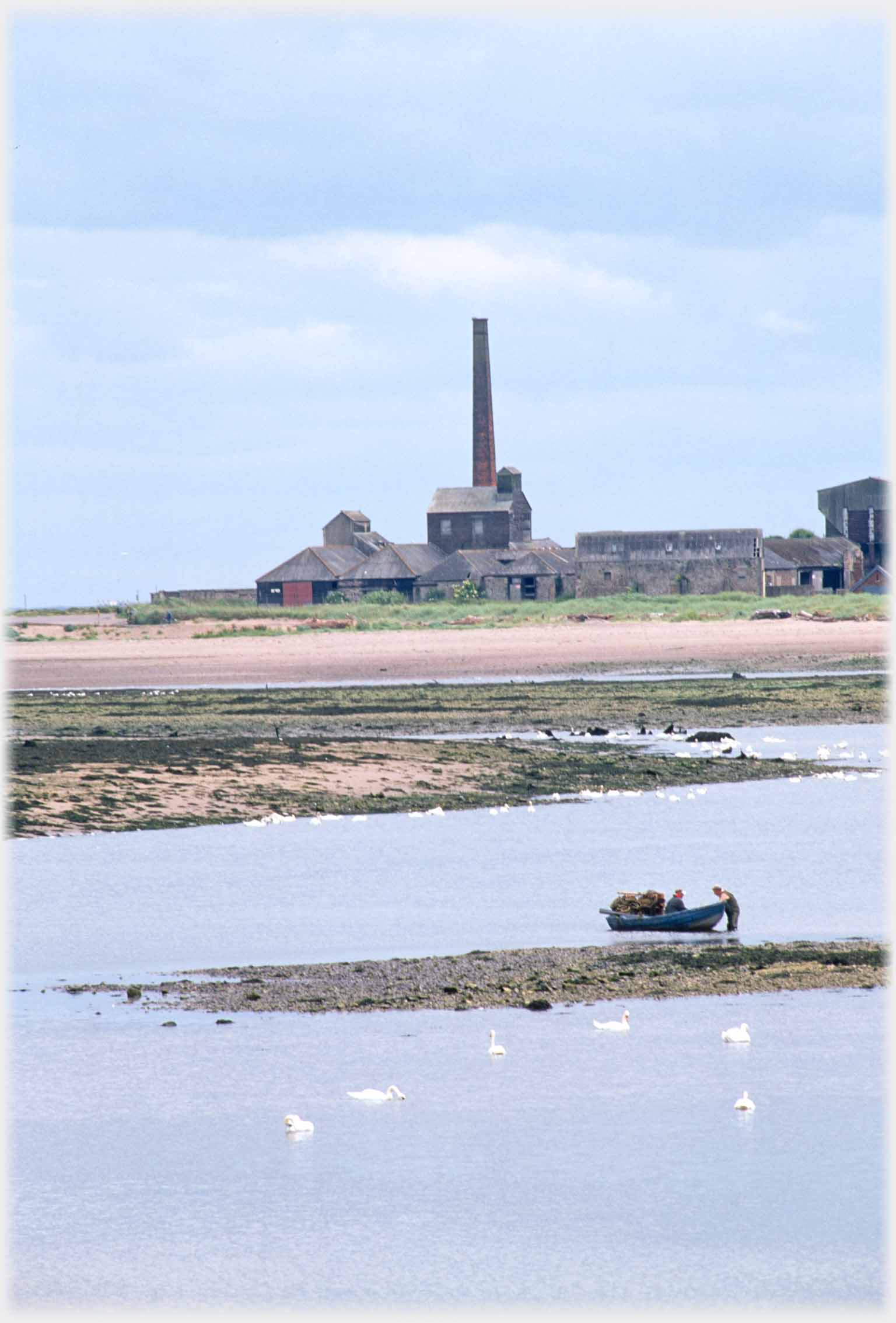 Factory with tower, small boat with men on water in foreground.